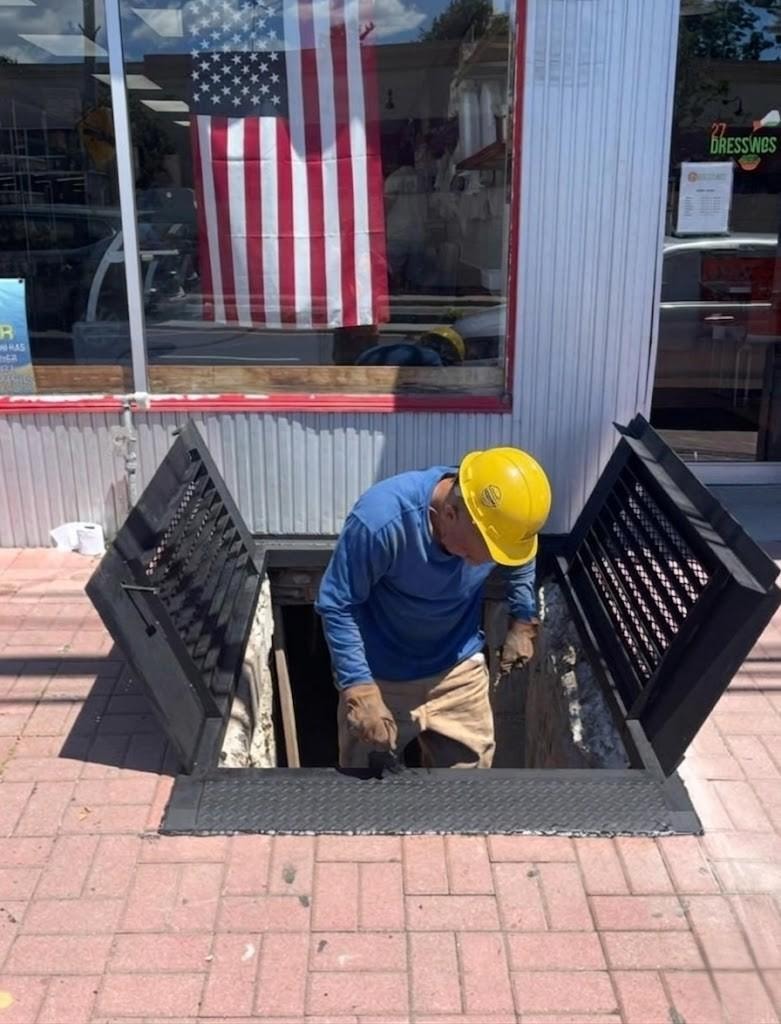 Worker in yellow hard hat descending into open diamond-plate sidewalk hatch